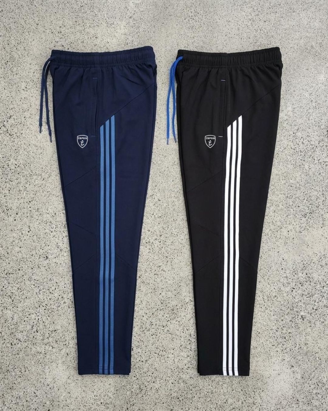 Two slim fit track pants with side stripes, drawstrings, and athletic logo on concrete background