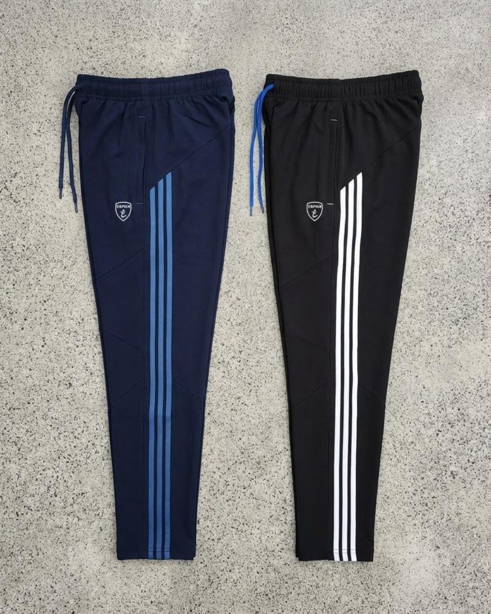 Two slim fit track pants with side stripes, drawstrings, and athletic logo on concrete background