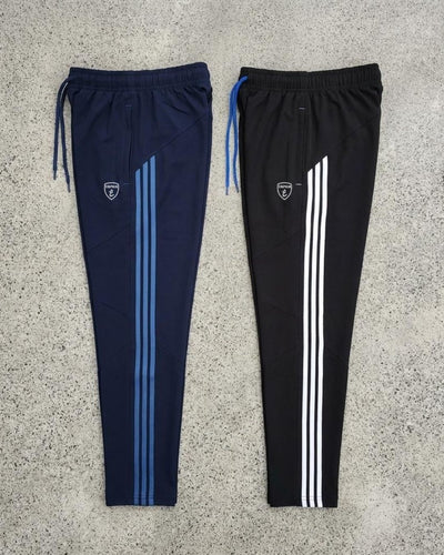 Two slim fit track pants with side stripes, drawstrings, and athletic logo on concrete background