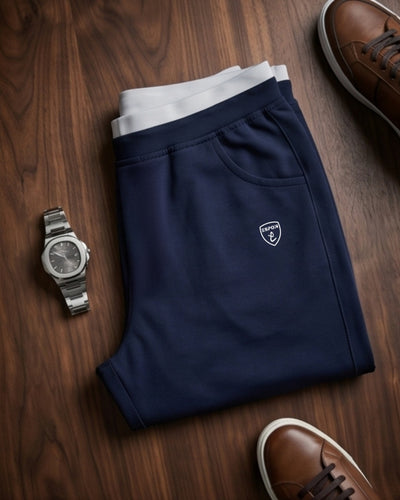 Navy blue athletic track pants with white waistband folded on wood, watch and brown sneakers nearby
