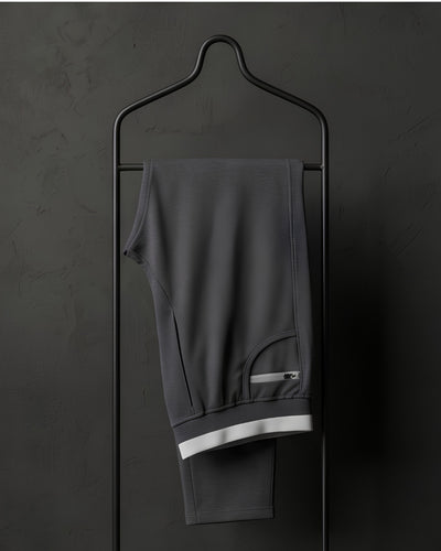 Dark grey track pants with white trim and zip pocket on black hanger against dark wall